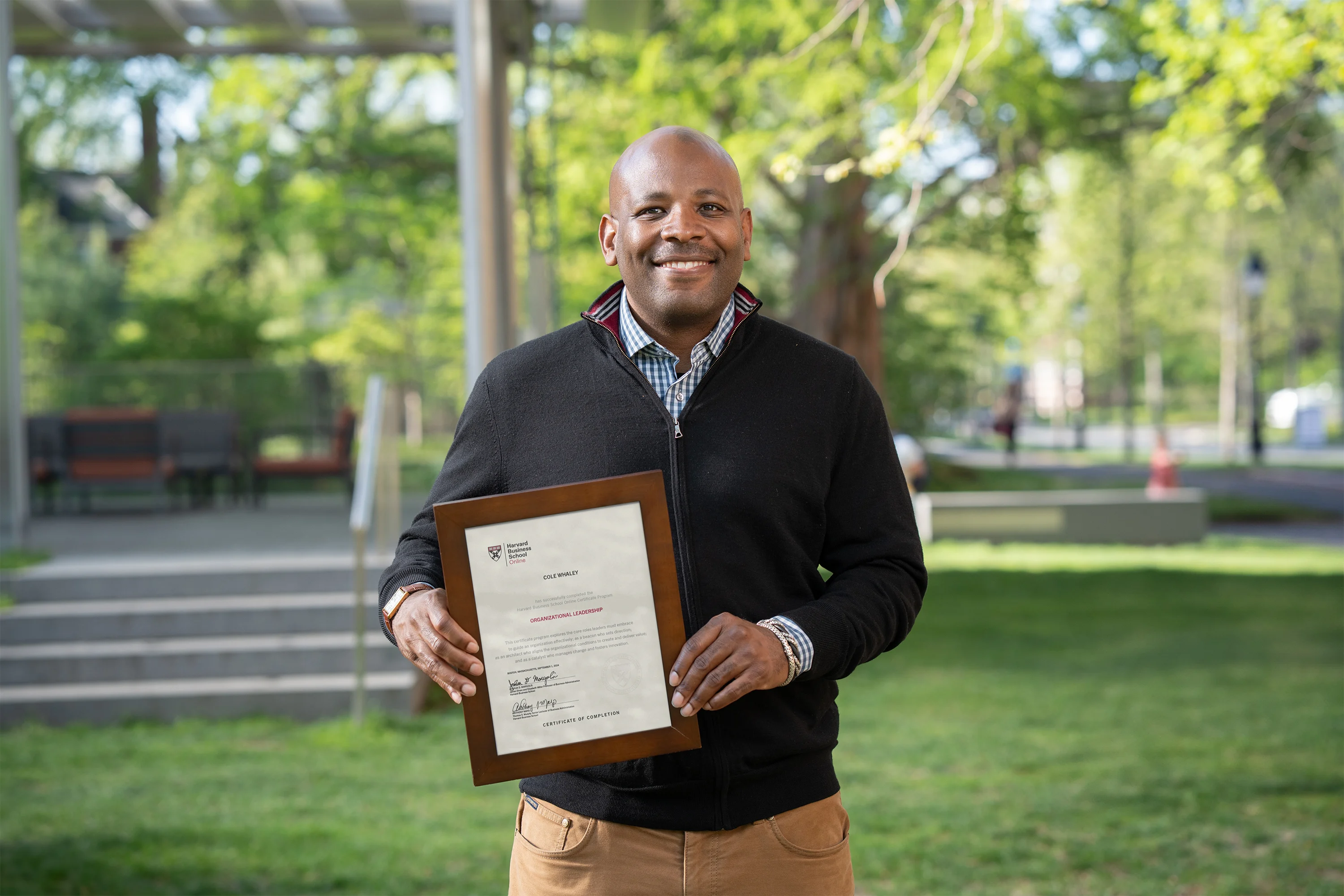 Person standing outdoors holding a framed Harvard Business School Online certificate and smiling at the camera.