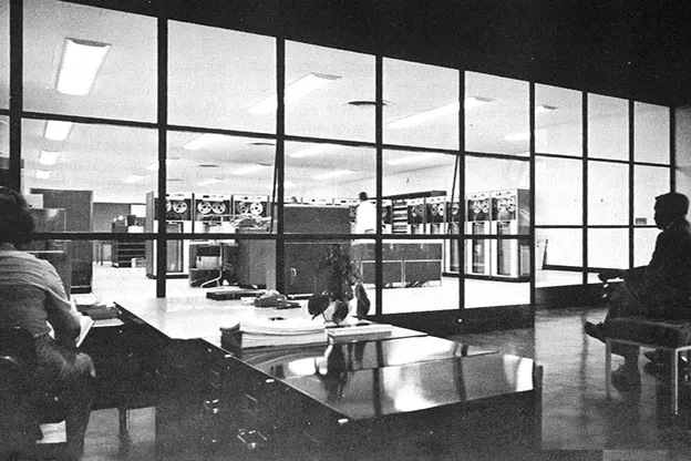 Two individuals sitting in a darkened room, one at a desk, one on a bench. Both are facing a lightened room with glass windows that holds many computers, circa 1963.