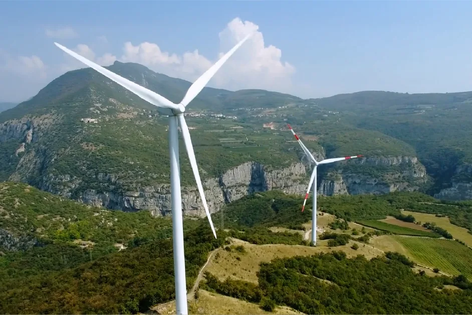 Two large wind turbines on a green hillside with rocky mountains in the background under a blue sky.