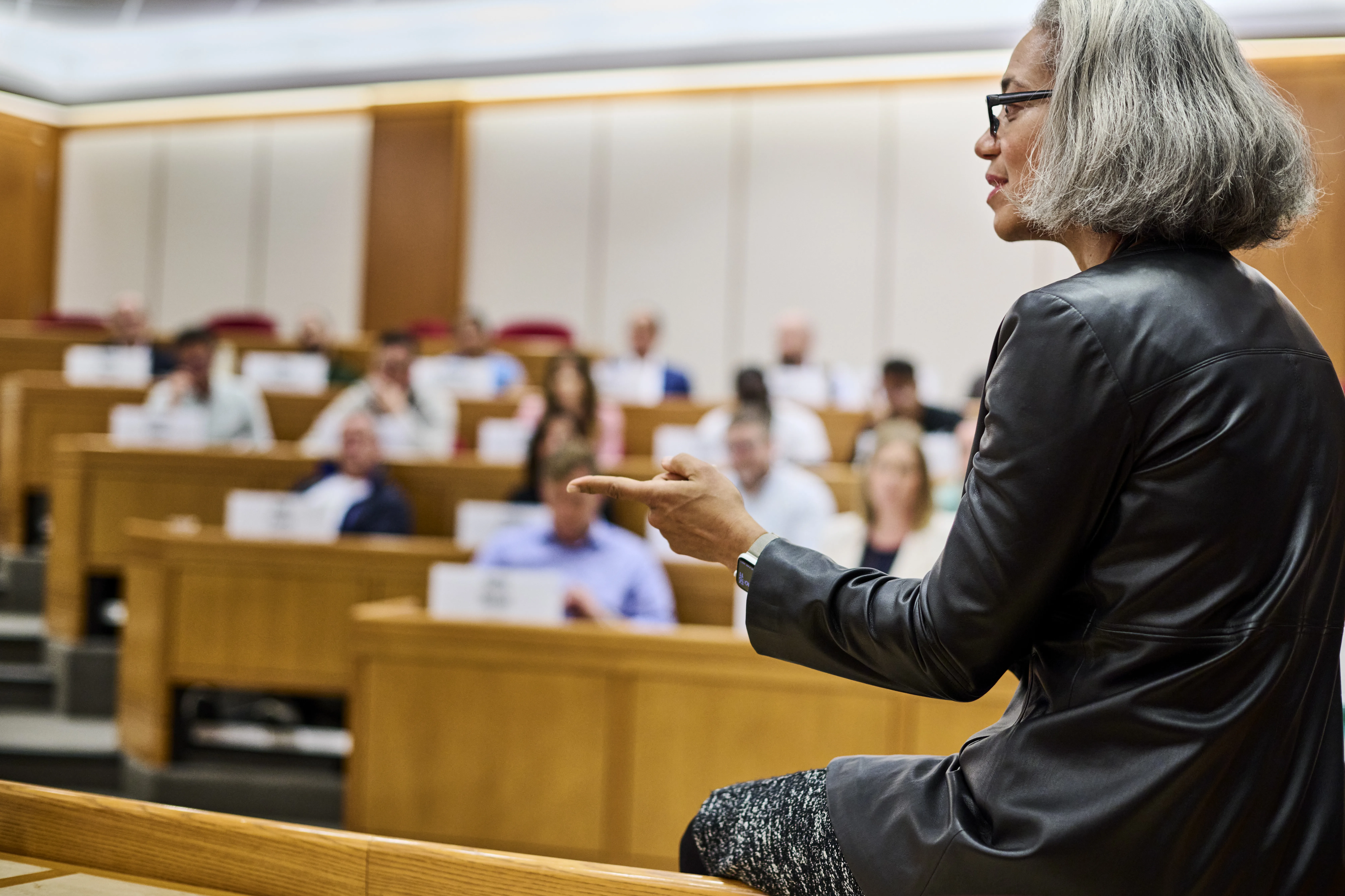 Instructor speaking to students in a lecture hall with tiered wooden desks and name placards.