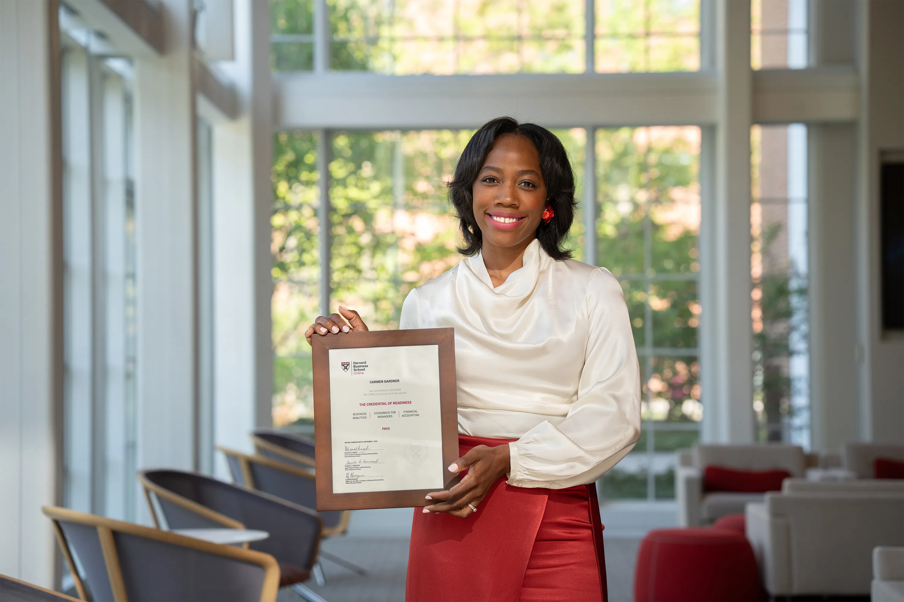 Individual in white blouse and red skirt smiling and holding a framed certificate in a sunlit lounge area.