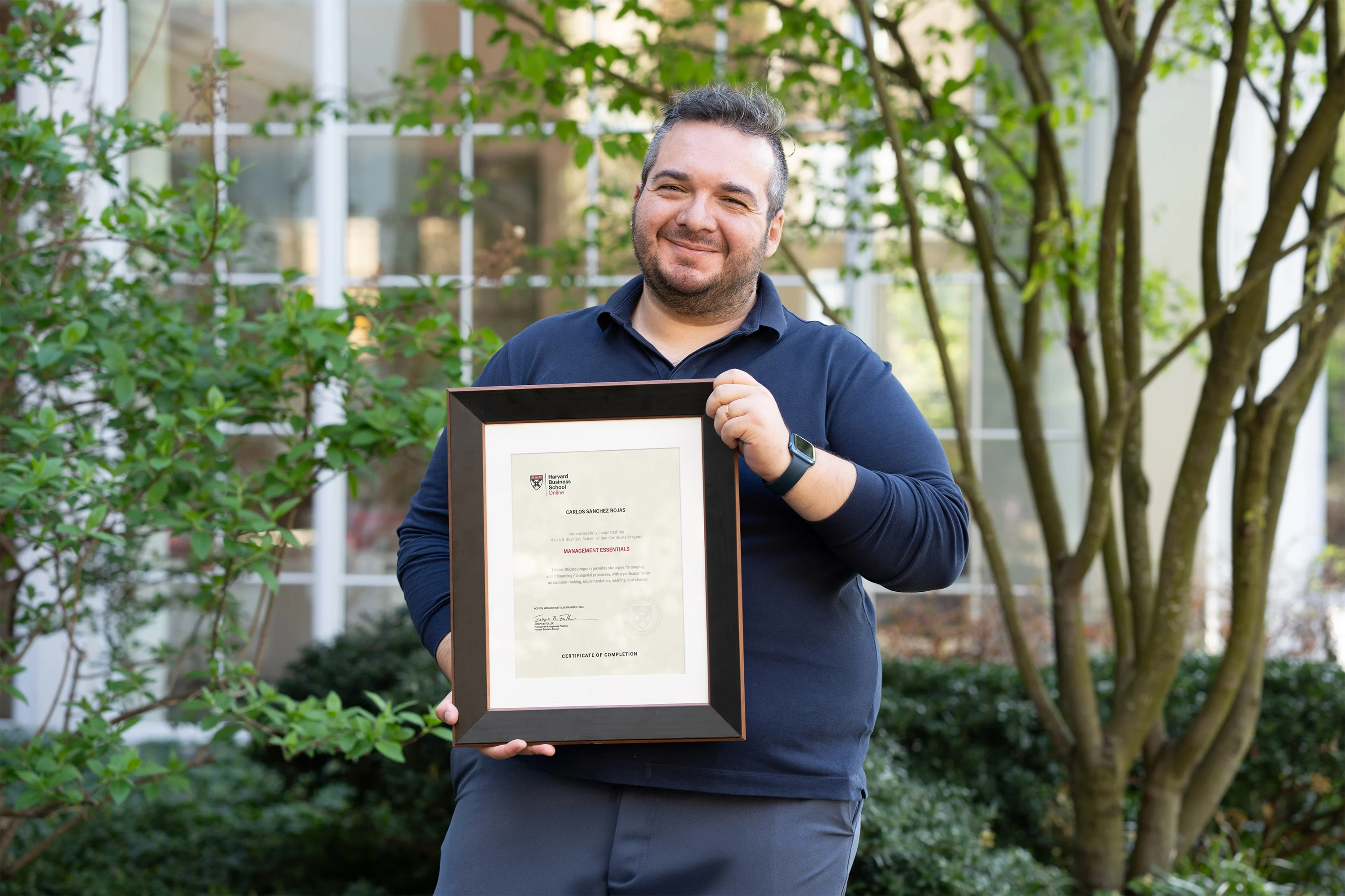 Individual holding a Harvard certificate of achievement, smiling outdoors near campus greenery.