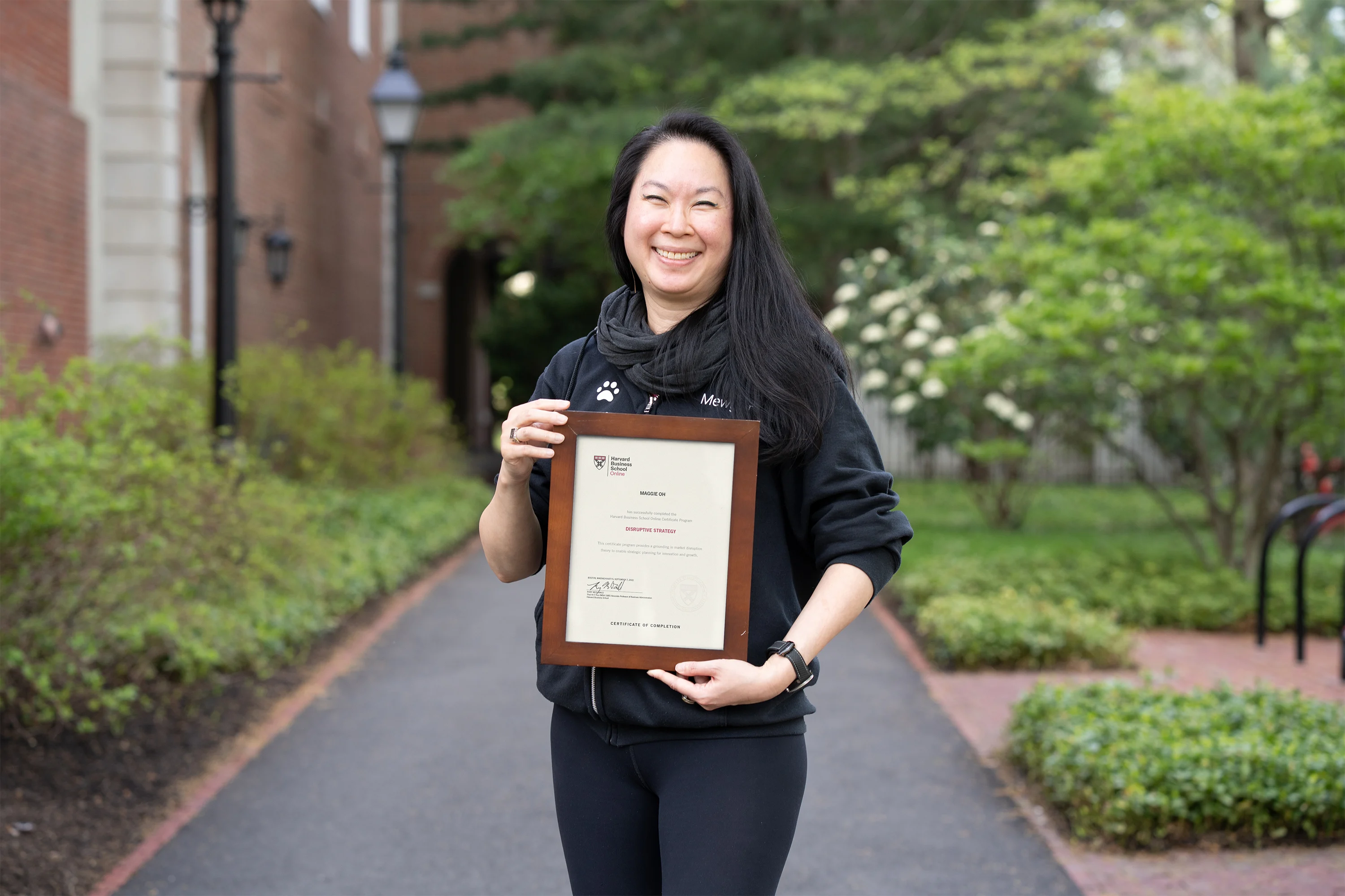 Person smiling and holding a framed certificate while standing on a brick pathway in a green campus setting.