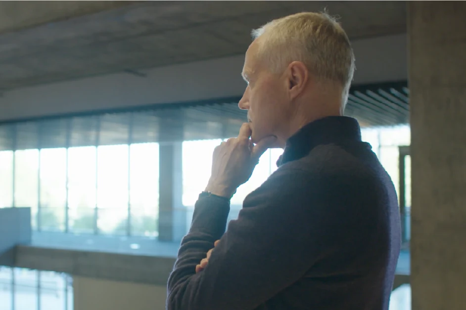 Person with short gray hair stands in a modern building, looking out thoughtfully with hand on chin.