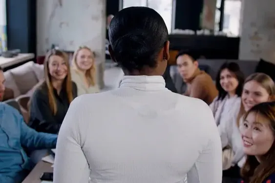 Person standing and speaking to a seated group of smiling individuals in a casual indoor setting.