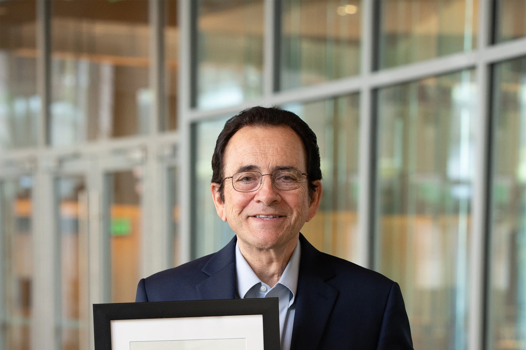 Individual in a dark suit smiling and holding a framed certificate in a modern glass-walled hallway.