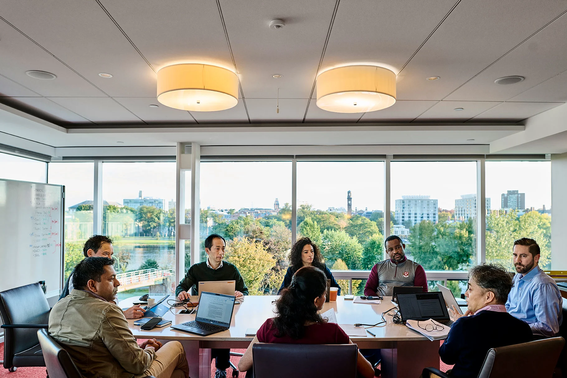 Professionals collaborate around a conference table in a bright meeting room overlooking the HBS campus landscape