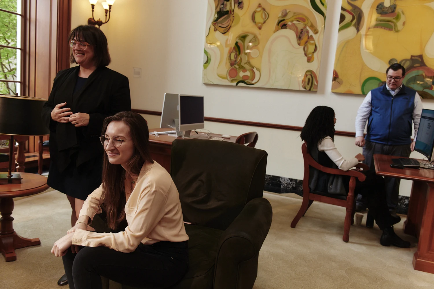 Librarians and students talking in the exchange lobby area seated on couches. In background librarian instructing student at a computer terminal.