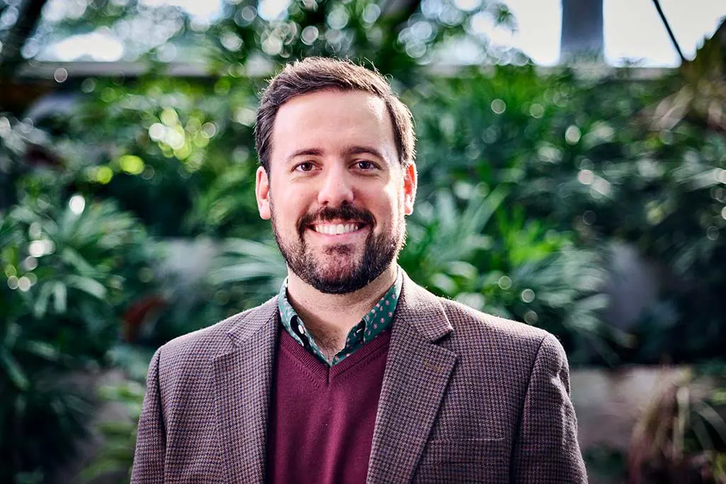 Portrait of an Executive Education participant in a plaid blazer with a green collared shirt and maroon sweater. Greenery surrounds him in the background.