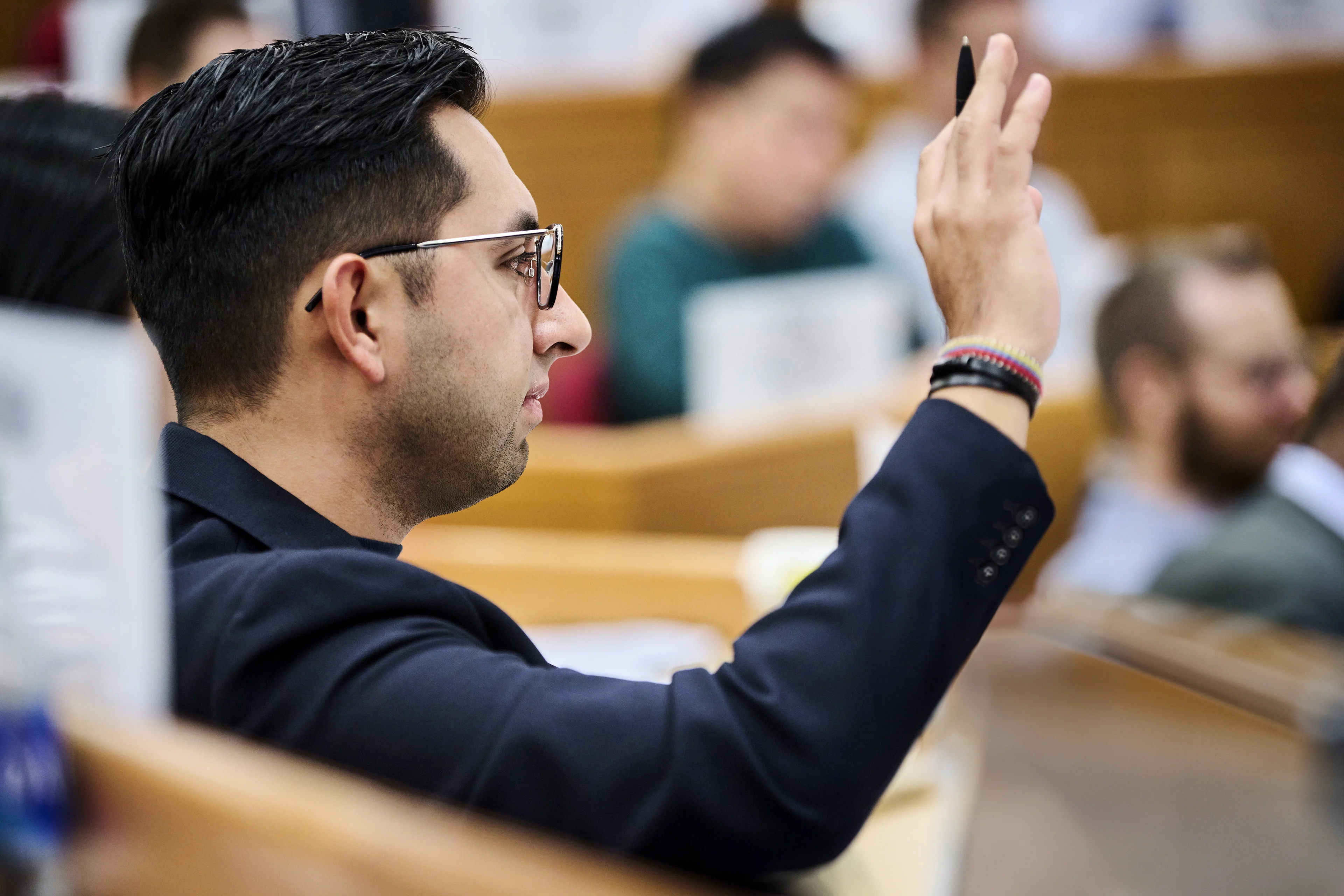 Male participant with black hair and glasses raising his hand in HBS classroom. 