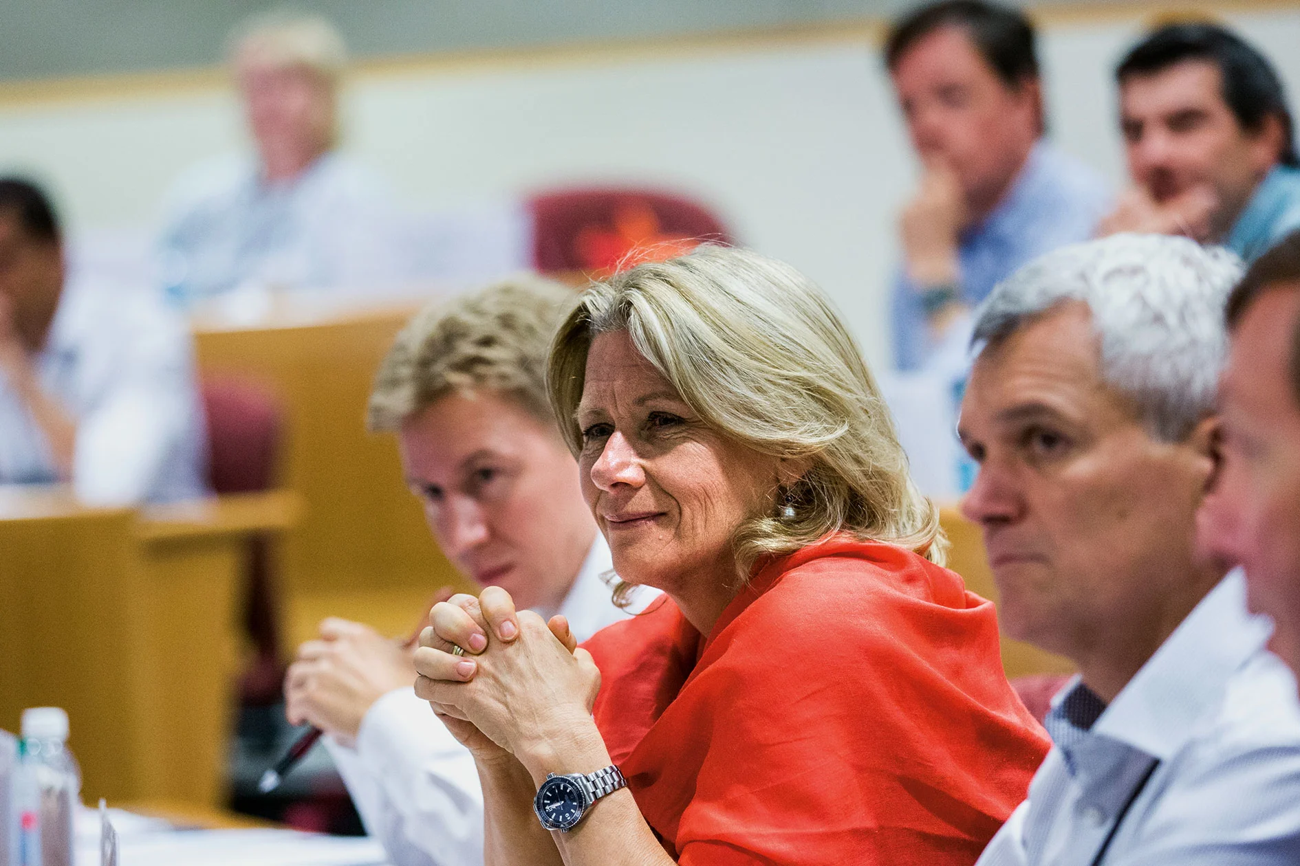 A group of executives attentively listening in a lecture hall, with a woman in a red shawl smiling