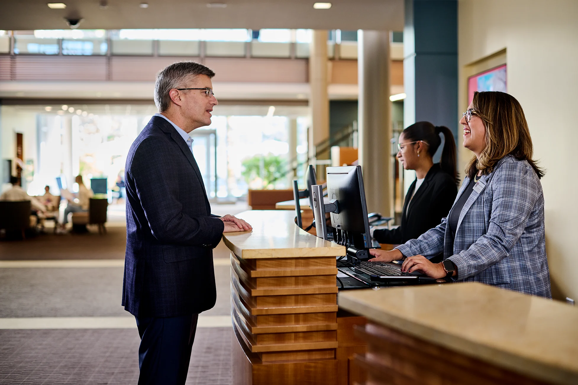 Man in black suit standing at a reception desk asking a question, two women behind the desk responding