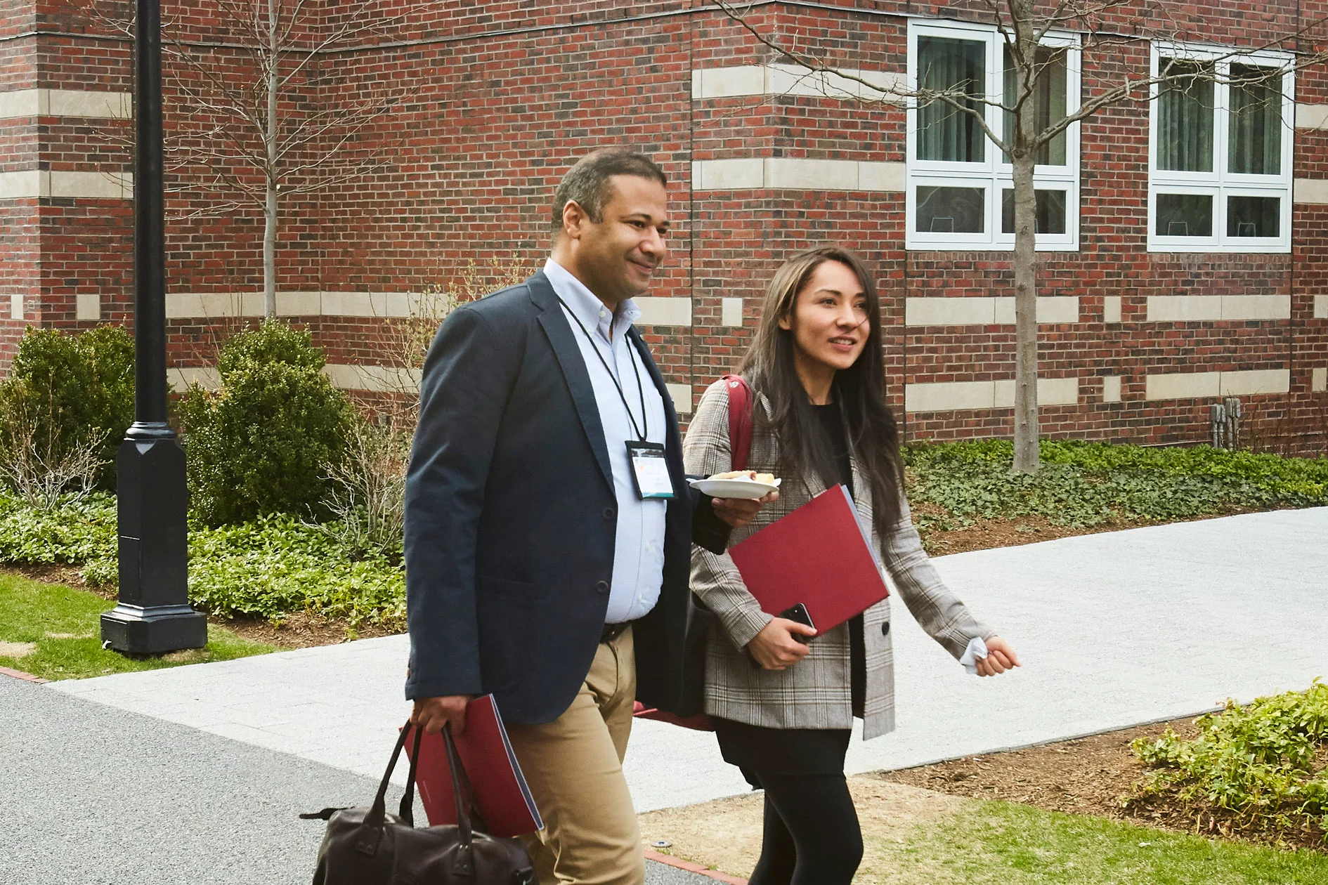 Two executives walk and chat on the HBS campus, carrying folders and bags, dressed in business casual attire