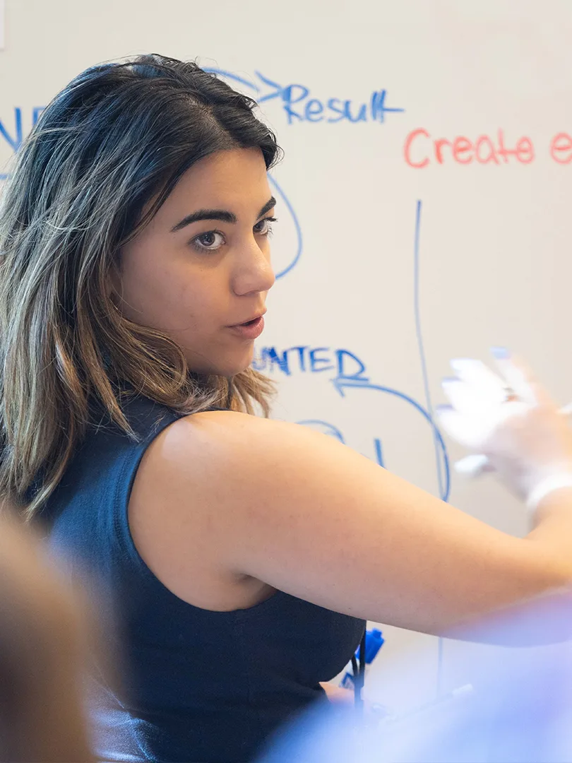 A Predoctoral Research Associate explain her writing on the whiteboard.