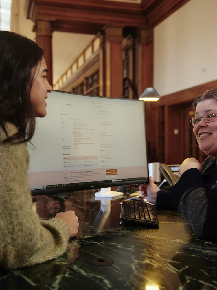 Library user and staff member interacting at the service desk of Baker Library.  Computer monitor screen and marble desk surface visible.