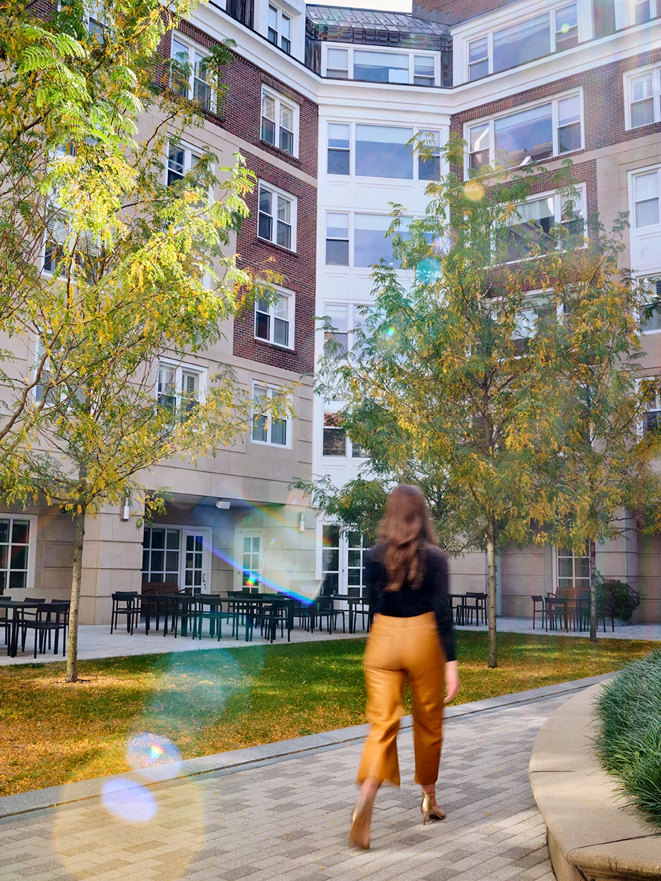 A woman in stylish attire walks through a sunlit McArthur courtyard surrounded by modern brick buildings and autumn tree