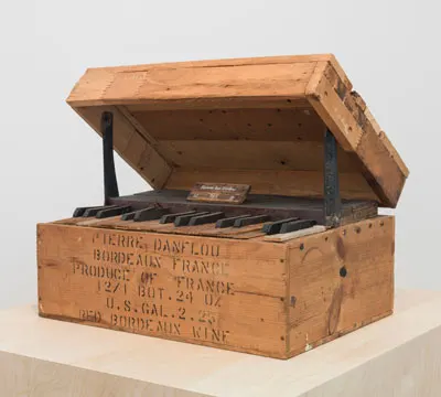 Wooden wine crate transformed into a small upright piano with black keys, lid propped open, and label on top.