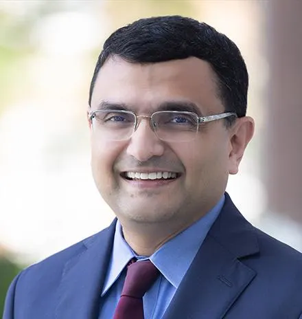 V.G. Narayanan wearing glasses, a navy suit, a light blue shirt, and a burgundy tie. Blurred outdoor background.