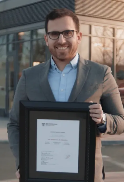 A man in a grey suit, blue shirt, and glasses smiles while holding a framed certificate