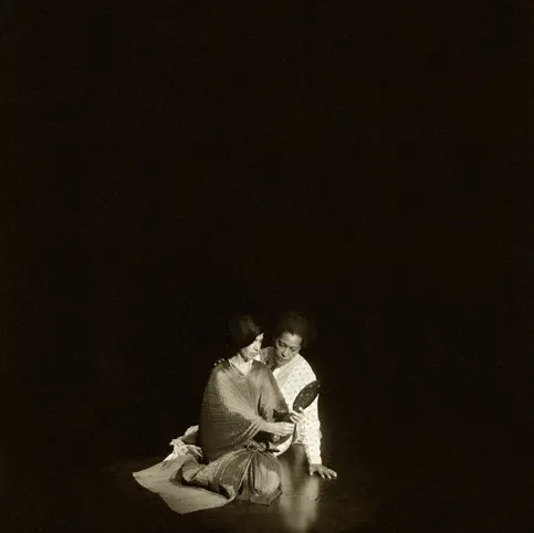 Two individuals sit closely together in darkness, softly lit, one holding a mirror, evoking intimacy and reflection.