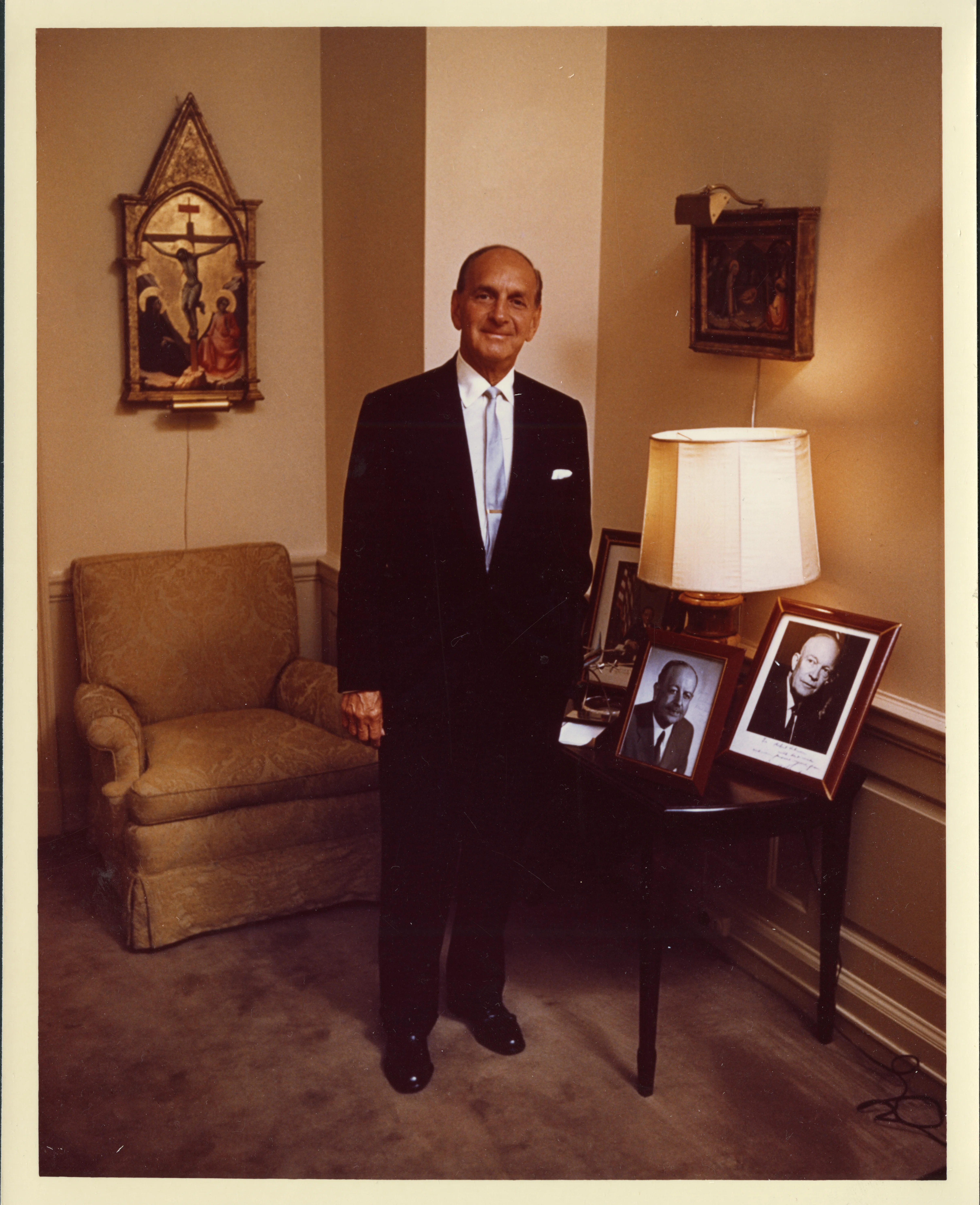 A man in a formal suit standining in a living room near a chair, art, and photos