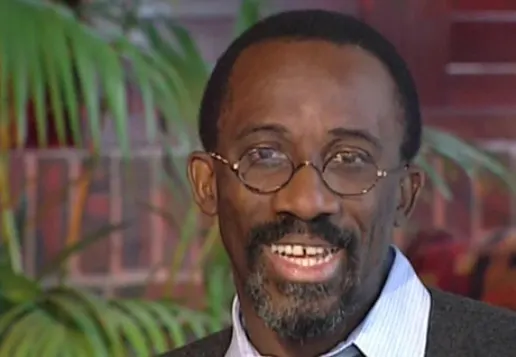 Smiling person with round glasses, short hair, and a goatee, wearing a light blue shirt and dark jacket indoors with plants behind them.