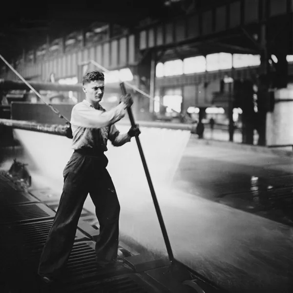 Factory worker uses a long rod to guide metal sheet through a water cooling process inside an industrial plant.