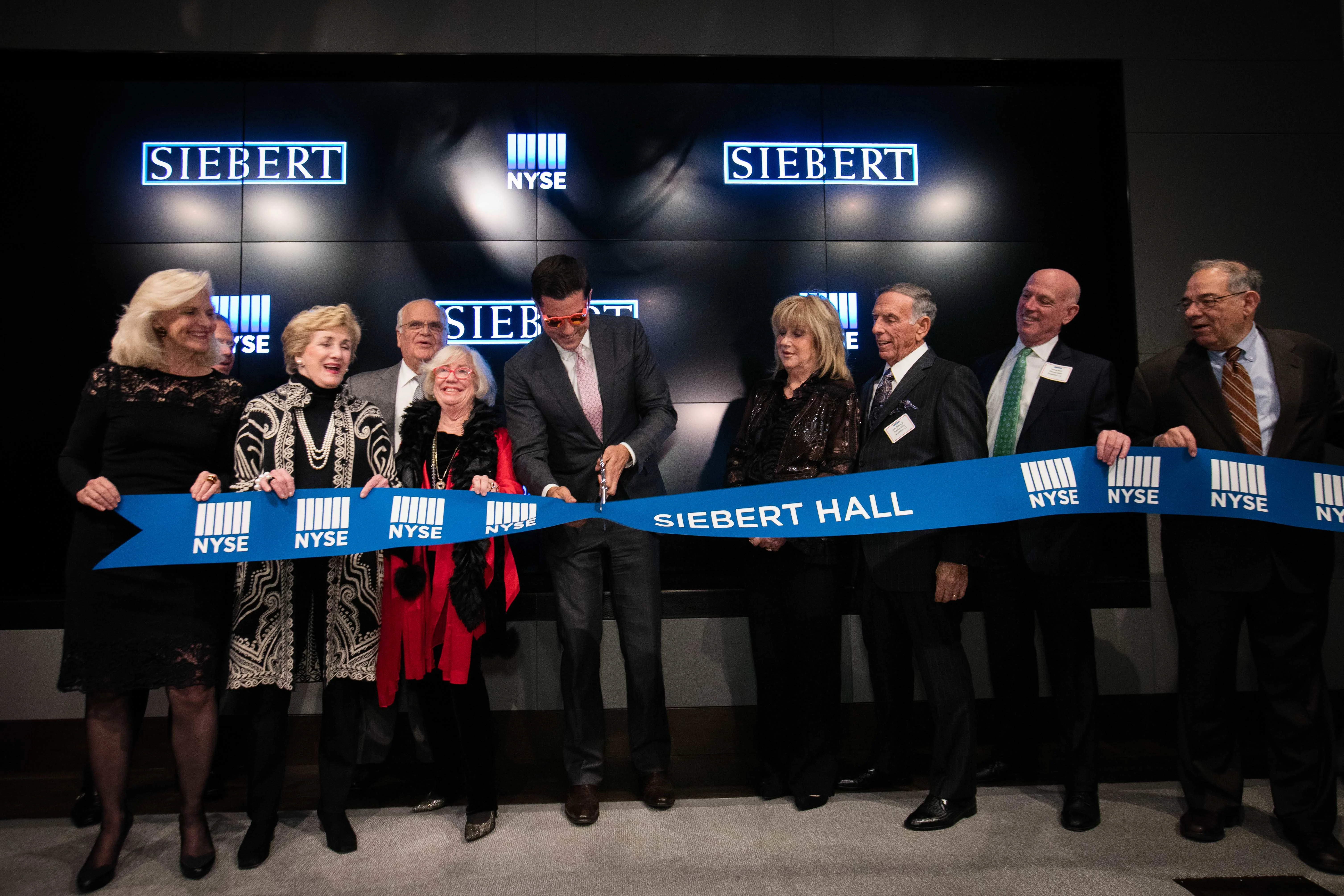 Group of people celebrate Siebert Hall ribbon-cutting at NYSE, with man in center cutting a blue ribbon.