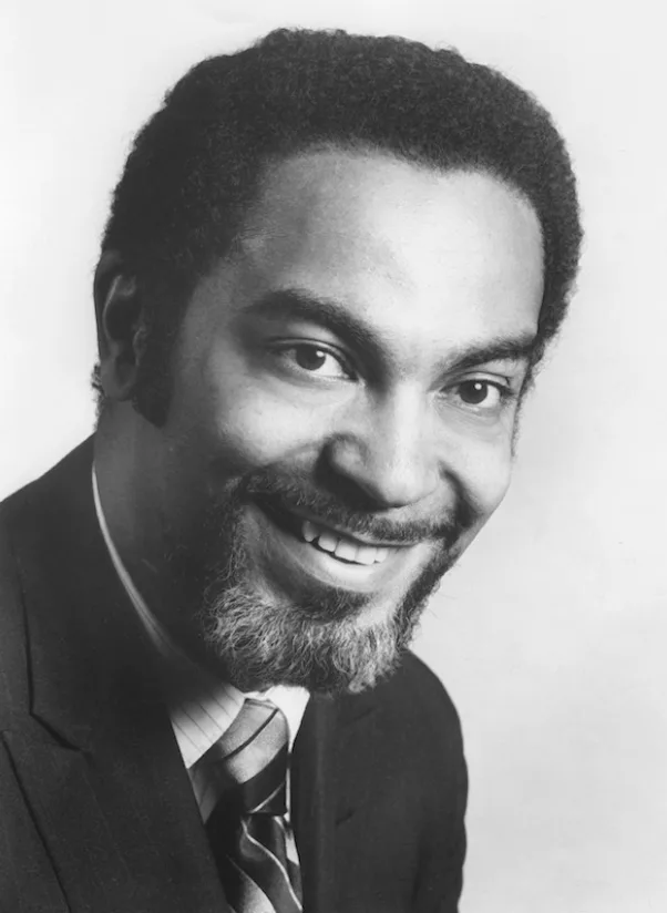 A Black man with a goatee and tie smiling.