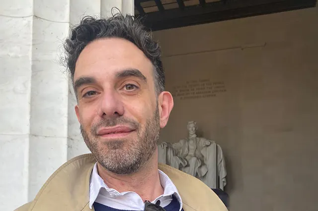 A white man with short curly dark hair and a slight beard stands in front of the Lincoln Momument.