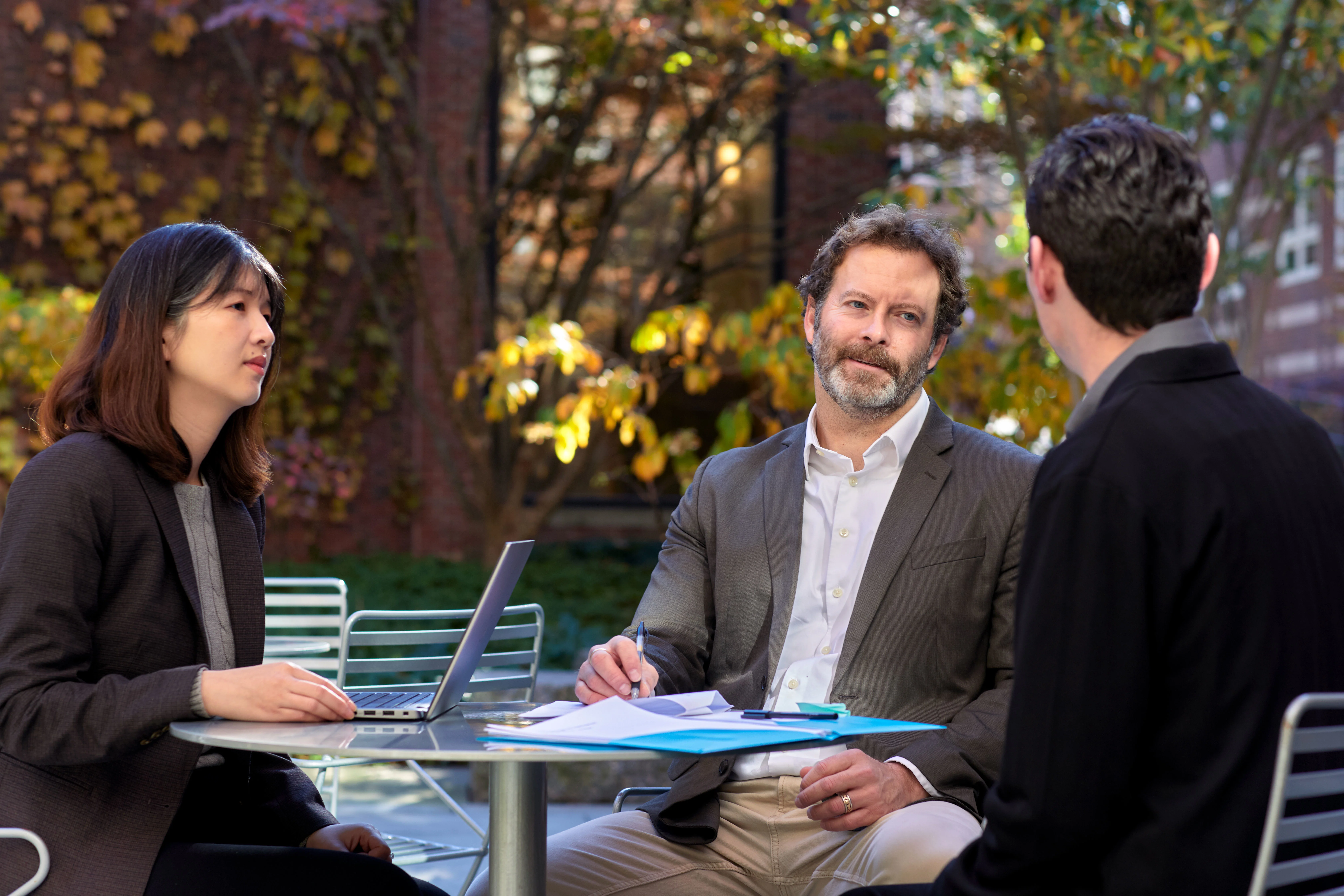 Three professionals meet around an outdoor table with a laptop and documents in a campus courtyard.