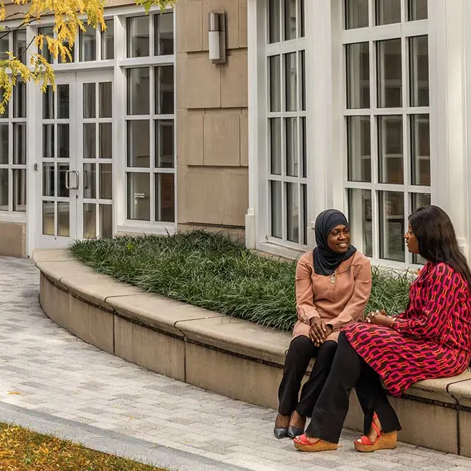 Two women of color sitting on a curved stone bench outside of Shad hall, engaged in a warm conversation.