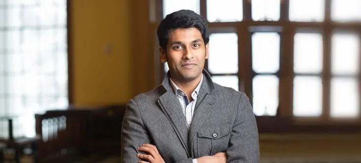 Person in a grey blazer stands with arms crossed in front of large window with grid-like panes.