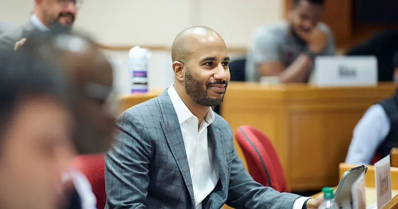 A professionally dressed male executive in a gray plaid suit sits in a HBS classroom, smiling and engaged in discussion