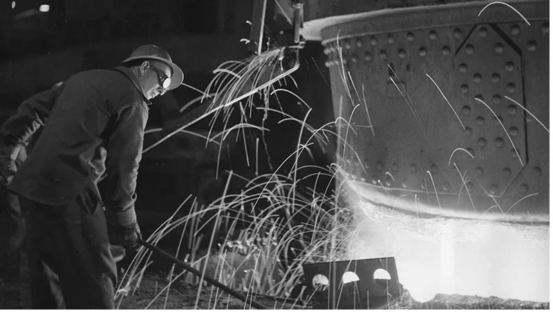 Worker in protective gear monitors molten metal pouring from a large furnace, with sparks flying.