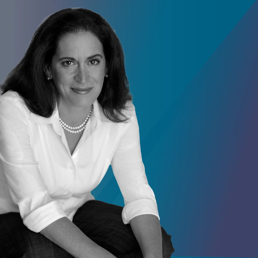 A woman with curly hair sits in a chair, facing the camera. One hand rests under her chin. She wears glasses on her head, a patterned blazer, and a light sweater. The background shows soft shapes in blue and orange. The woman is in black and white.