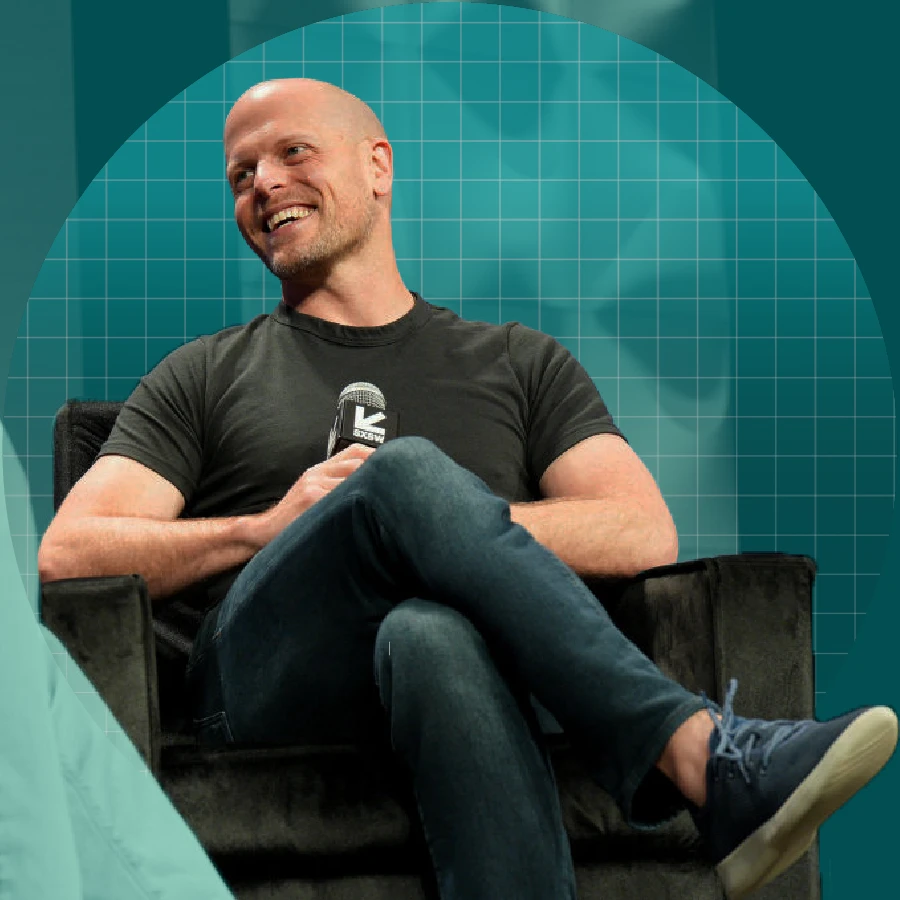 Tim Ferriss sitting on a dark armchair, holding a microphone. He wears a fitted dark T-shirt, blue jeans, and light-colored lace-up shoes. His legs are crossed, and he's turned slightly. Behind him is a teal circular backdrop with a grid pattern.