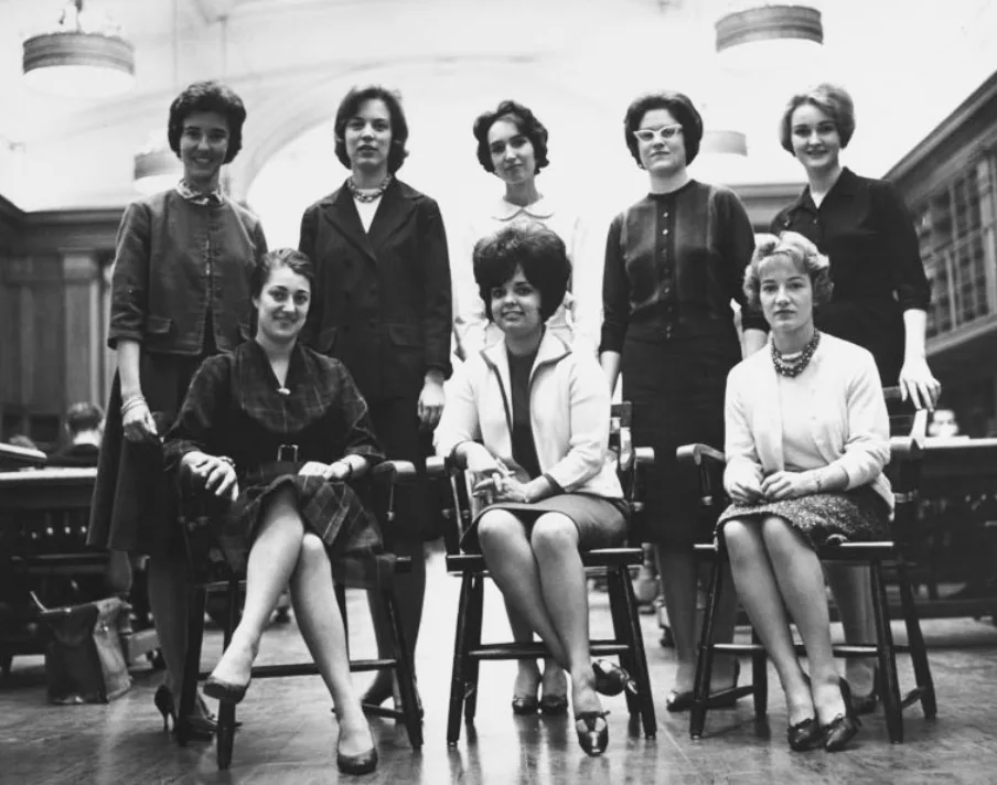 Eight women in mid-20th-century attire pose in a library. Three sit in front, five stand behind. The setting includes bookshelves and large hanging lights. Some wear jewelry or glasses.