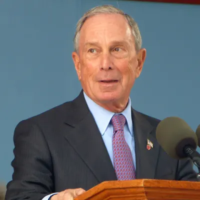 Person in a suit and red patterned tie speaking at a podium with microphones, against a blue background.