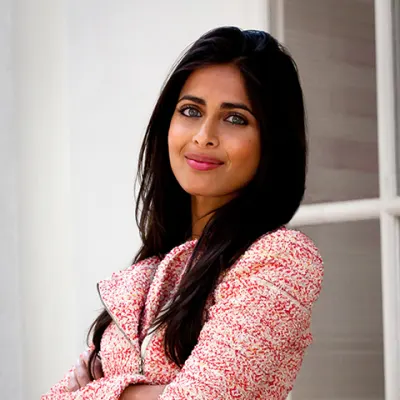 Person with long dark hair wearing a textured pink blazer, standing in front of a white wall and window.
