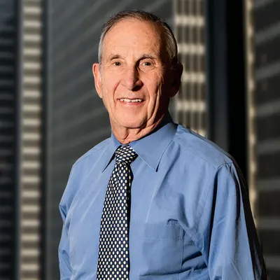 Person wearing a blue dress shirt and patterned tie, standing in front of tall office buildings in sunlight.