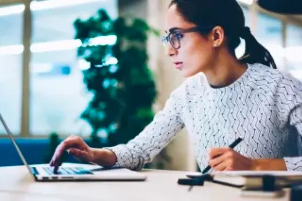A woman in glasses sits at a table, one hand holding a pen, the other typing on her laptop