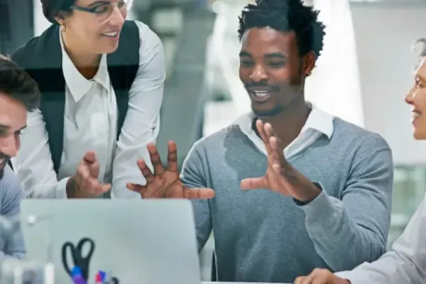 A man sits surrounded by coworkers, gesturing while speaking in front of his laptop screen