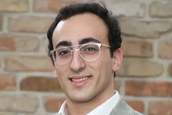 Person with short dark hair and glasses wearing a light gray blazer and white shirt, smiling in front of a brick wall.