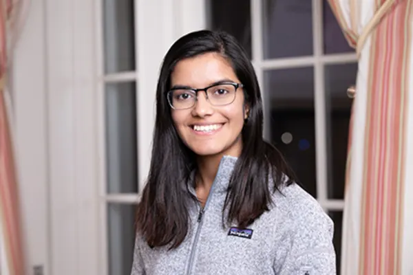 A young woman with glasses wearing a gray sweater, looking directly at the camera with a friendly expression.
