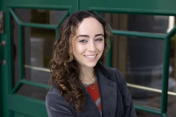 A woman with long curly hair stands in front of a vibrant green door, smiling and looking towards the camera.