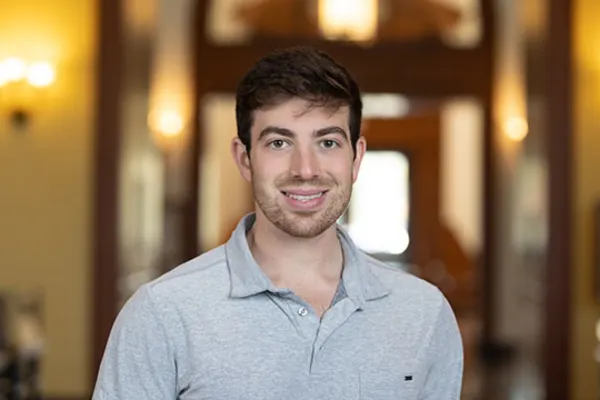 A man in a gray shirt smiles warmly at the camera, exuding a friendly and approachable demeanor.