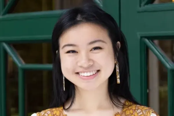 Person with long dark hair wearing floral-patterned clothing and dangling earrings, smiling in front of green doors.