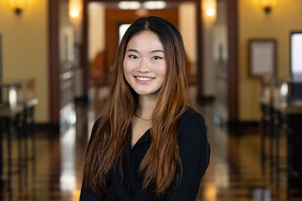 A young woman wearing a black shirt stands in a hallway, looking confidently ahead.