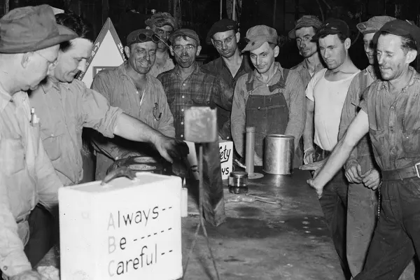 Group of factory workers gather around a table with a safety sign that reads "Always Be Careful."